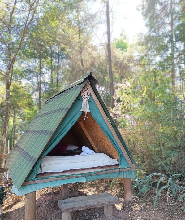 A small, triangular wooden tent with a green roof, a sleeping area with a white mattress, and a decorative hanging ornament.