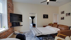 Een moderne woonkamer met een flatscreen televisie aan een bakstenen muur, een bruine leren bank, een bed met een witte deken en een vloerkleed met dessin.