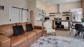 Een moderne woonkamer met een bruine leren bank, een witte fauteuil, en een eethoek met een houten tafel en stoelen. Achtergrond toont een keuken met witte kastjes en een roestvrijstalen koelkast.