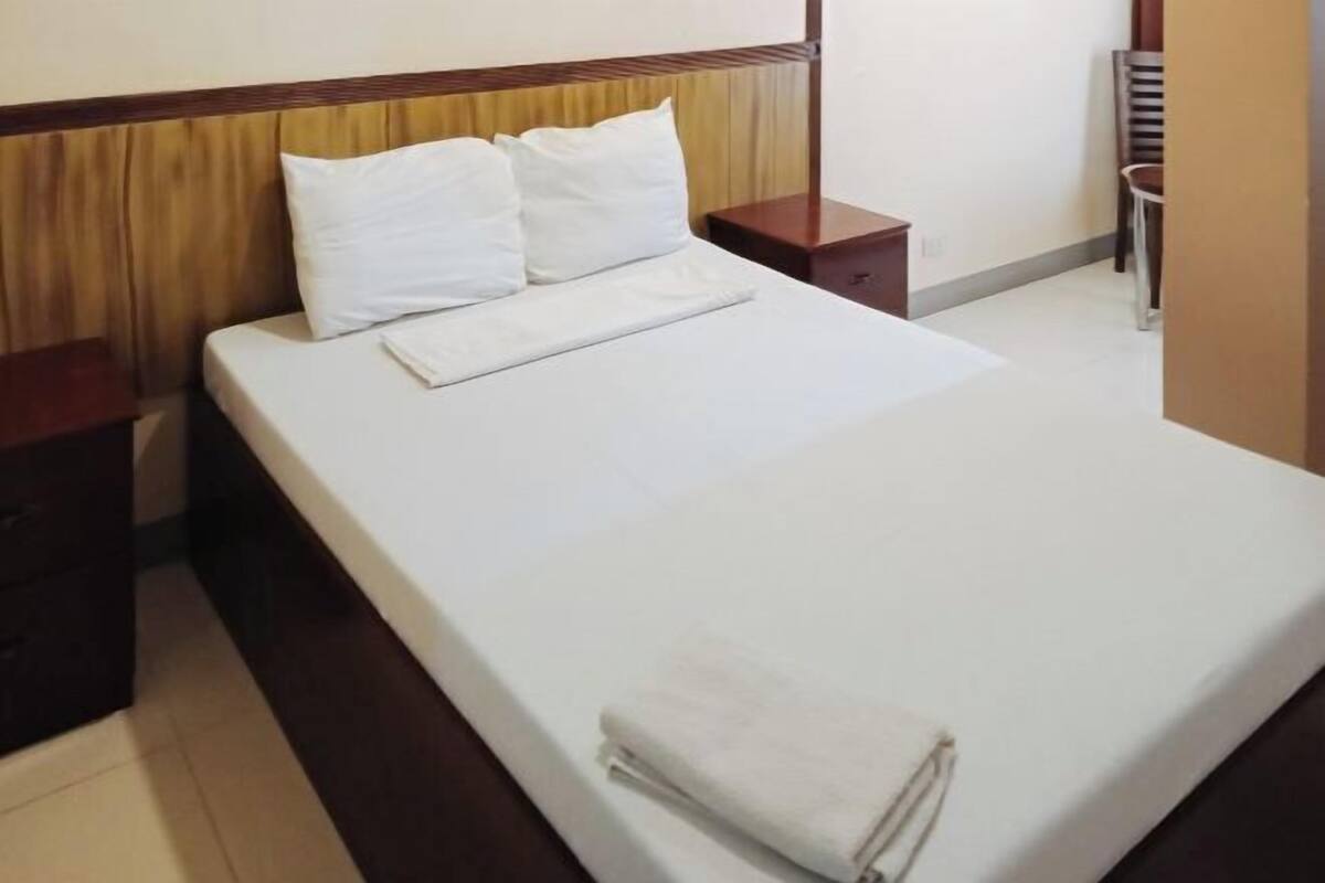 A neatly made bed with white linens and pillows, a wooden headboard, a small wooden nightstand, and a chair in the corner.