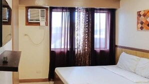 Una habitación de hotel con cama, una mesita, un equipo de aire acondicionado y una ventana con cortinas.