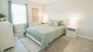 Un dormitorio con cama, mesitas de noche, ventilador de techo, ventana con cortinas y una lámpara de pared.