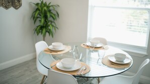 Una mesa redonda de vidrio con sillas blancas, dispuesta con platos, fuentes y cubiertos blancos. Un mantel tejido se coloca debajo de cada plato.