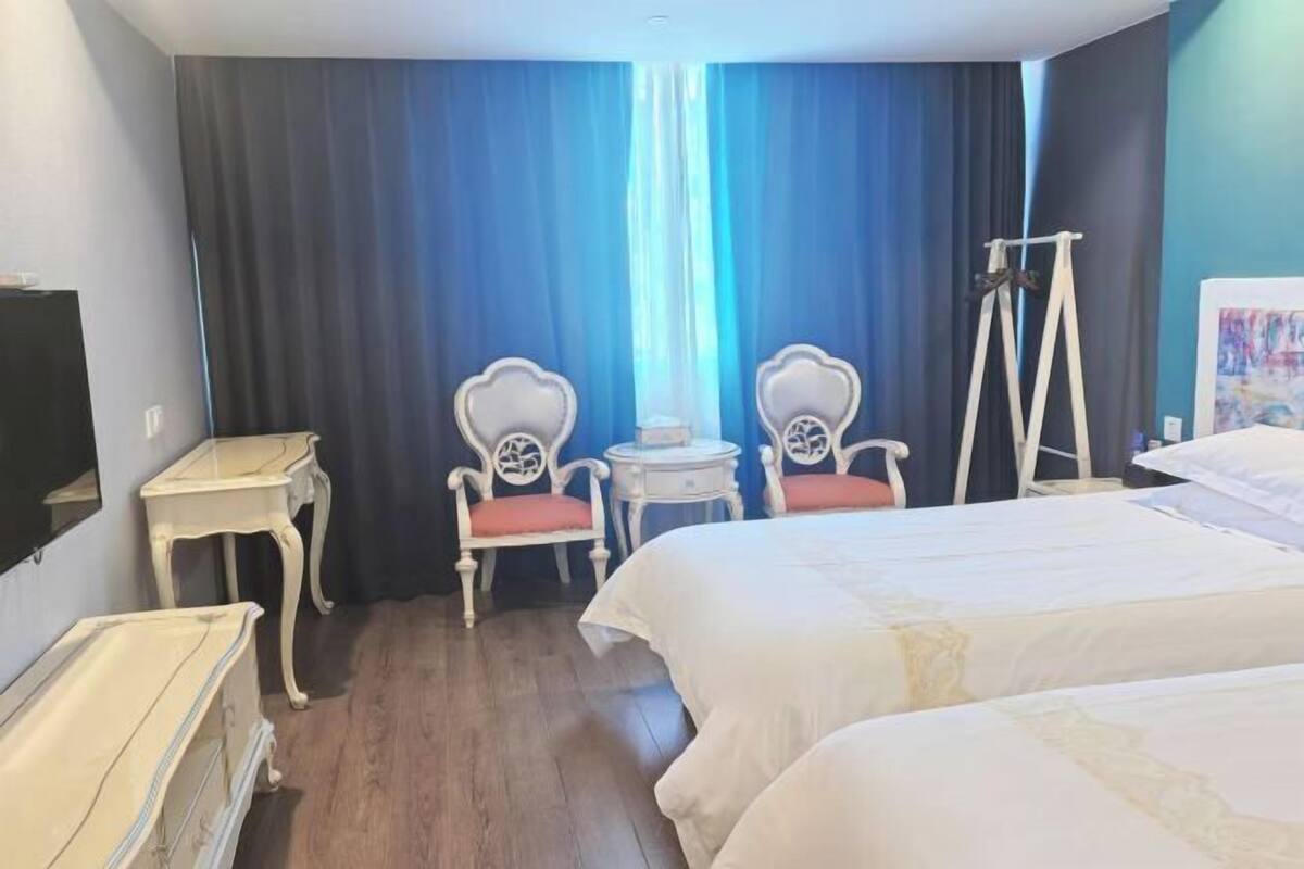 Camera d'albergo con due letti, un tavolino, due sedie, una televisione e una finestra con tende.