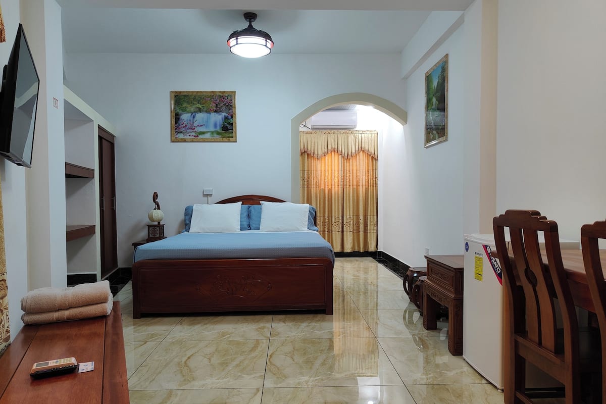 A bedroom with a bed, a desk, a chair, a TV, a painting, and a ceiling fan.