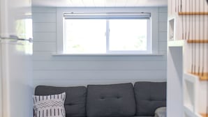 Un espacio habitable compacto con un sofá seccional gris, una ventana con persianas y piso de madera.