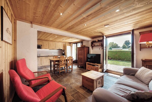 A spacious living area with a wooden ceiling, a dining table with chairs, a sofa, and a television.