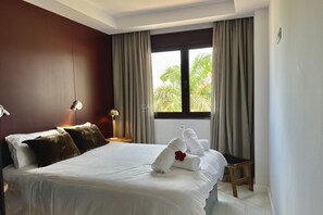 A hotel room with a bed, a nightstand, a lamp, a window with curtains, and a view of palm trees. - Luxury house with pool, jacuzzi, and views of the sea and golf course. (Santa Cruz de Tenerife)