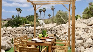 Une pergola en bois installée à l’extérieur, avec une table dressée pour les repas, entourée de rochers et de palmiers.