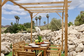 Una pérgola de madera al aire libre con una mesa dispuesta para comer, rodeada de rocas y palmeras.