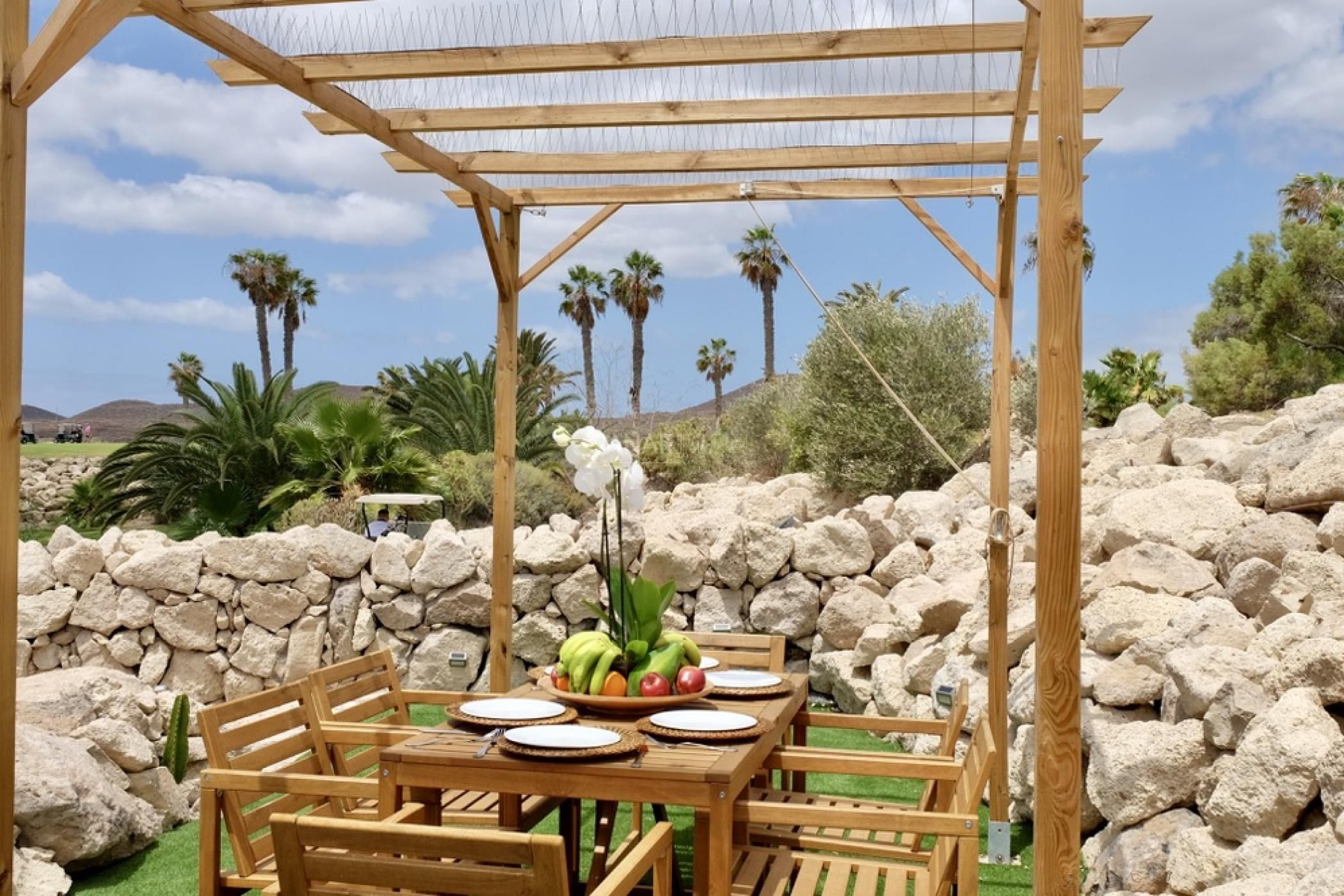 An outdoor wooden pergola with a table set for dining, surrounded by rocks and palm trees.