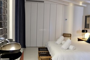 Una habitación de hotel con una cama, un taburete de madera, un calentador fijado a la pared, una mesita de noche con una lámpara y una ventana con cortina.