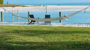 A hammock between two chairs on a wooden deck overlooking a beach and ocean.