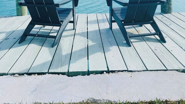 Two Adirondack chairs on a wooden deck overlooking a beach with clear blue water and a sandy shore.
