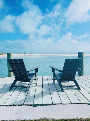Deux fauteuils Adirondack sont placés sur une terrasse en bois, d’où l’on a une vue sur une plage aux eaux d’un bleu limpide et au rivage sablonneux.