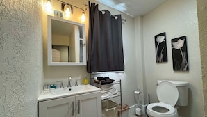 Une salle de bain avec un lavabo blanc, un miroir et des toilettes. Deux tableaux encadrés sont accrochés au mur, ainsi qu’un porte-serviettes et une étagère contenant des articles de toilette.