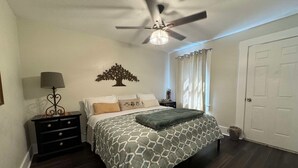 Une chambre à coucher comprenant un lit, une table de chevet, une lampe, un ventilateur de plafond, une fenêtre avec des rideaux et une porte.
