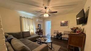 Un salon avec un canapé, une table basse et un ventilateur de plafond. Un téléviseur est fixé au mur, et une petite table est décorée de divers objets.