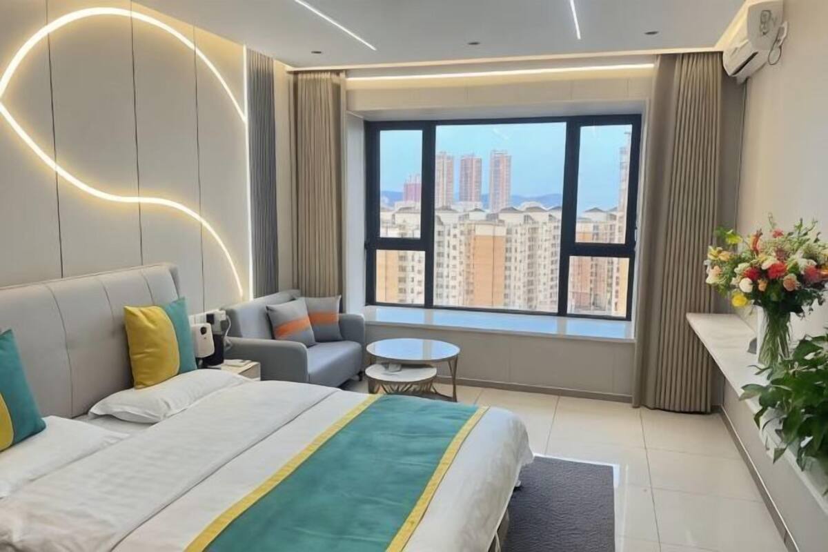 A modern hotel room with a large bed, a sofa, a small round table, and a view of tall buildings.