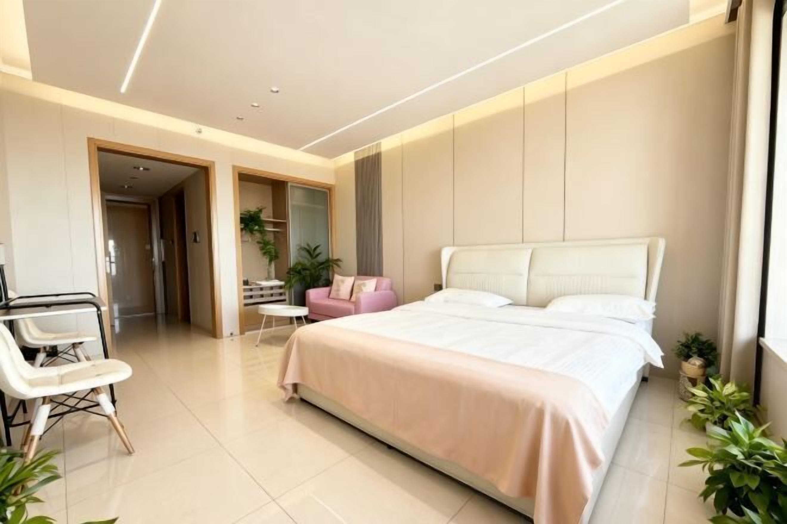 A modern bedroom with a large bed, a desk, and a chair. There are plants and a pink chair.