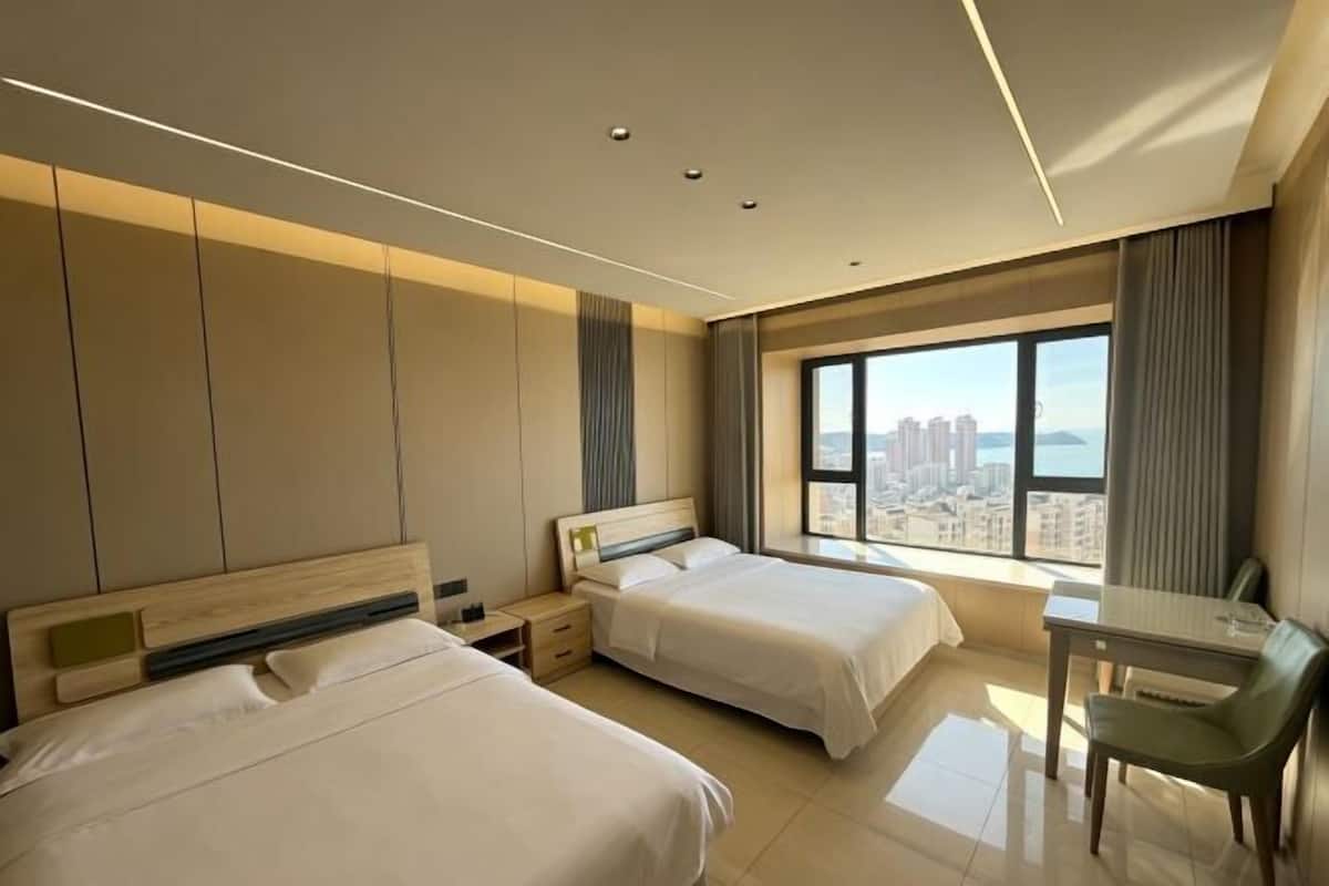 A hotel room with two beds, a desk, and a large window with a city view.