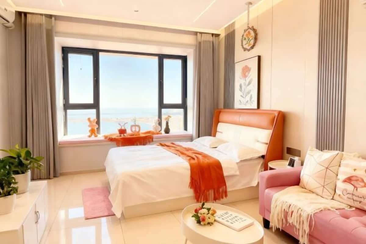 A modern bedroom with a large bed, a pink sofa, a view of the sea, and a window with curtains.