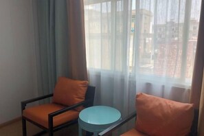Una habitación con dos sillas y una pequeña mesa redonda, una ventana con cortinas traslúcidas y vistas a edificios.