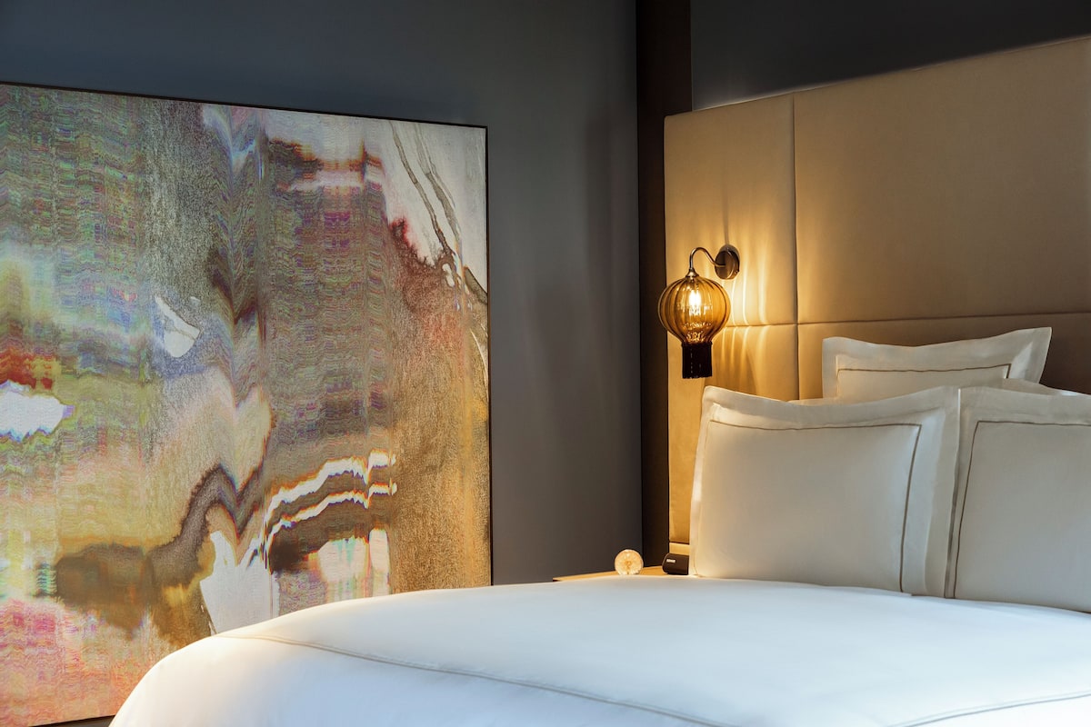 A modern bedroom with a large abstract painting, a plush bed, and a wall-mounted light fixture.