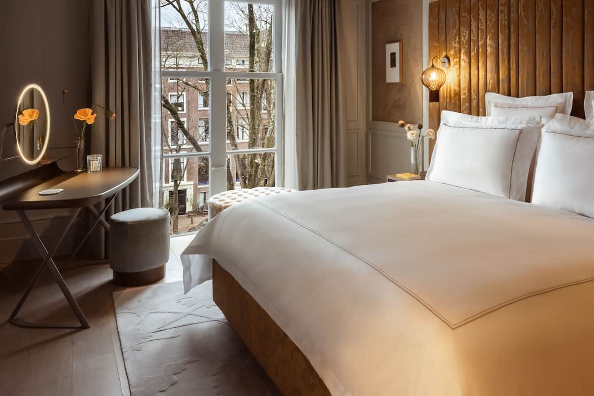 A hotel room with a large bed, a bedside table with a mirror, a small stool, and a view of the outside through a window.