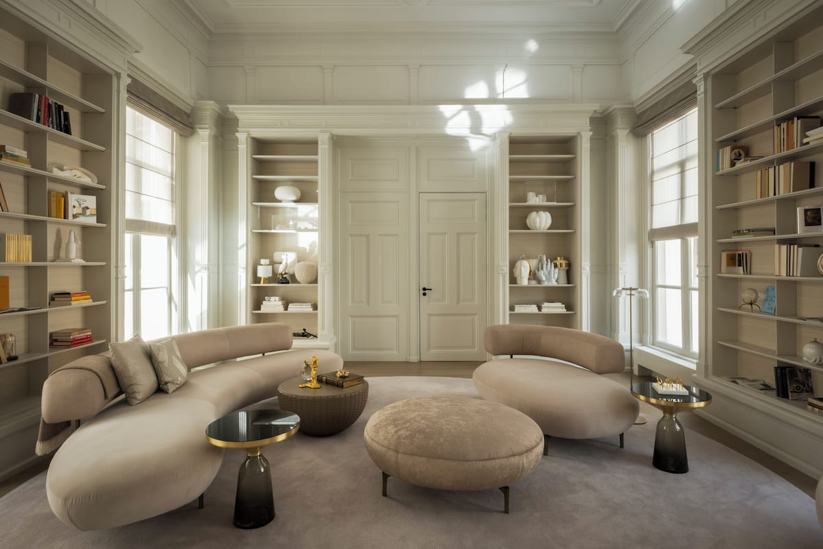 A spacious living room with a large L-shaped sofa, a round ottoman, and a coffee table. There are bookshelves on either side of the room, filled with books and decorative items.