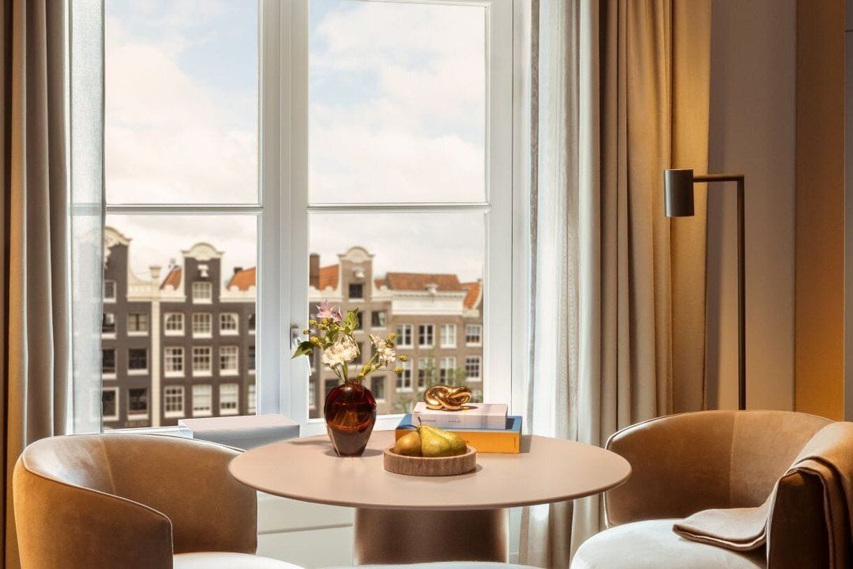Ein Zimmer mit einem runden Tisch, zwei Stühlen, einer Blumenvase, einer Obstschale und Blick auf Gebäude durch große Fenster.