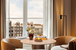 A room with a round table, two chairs, a vase with flowers, a bowl of fruit, and a view of buildings through large windows. - Rosewood Amsterdam (Amsterdam)