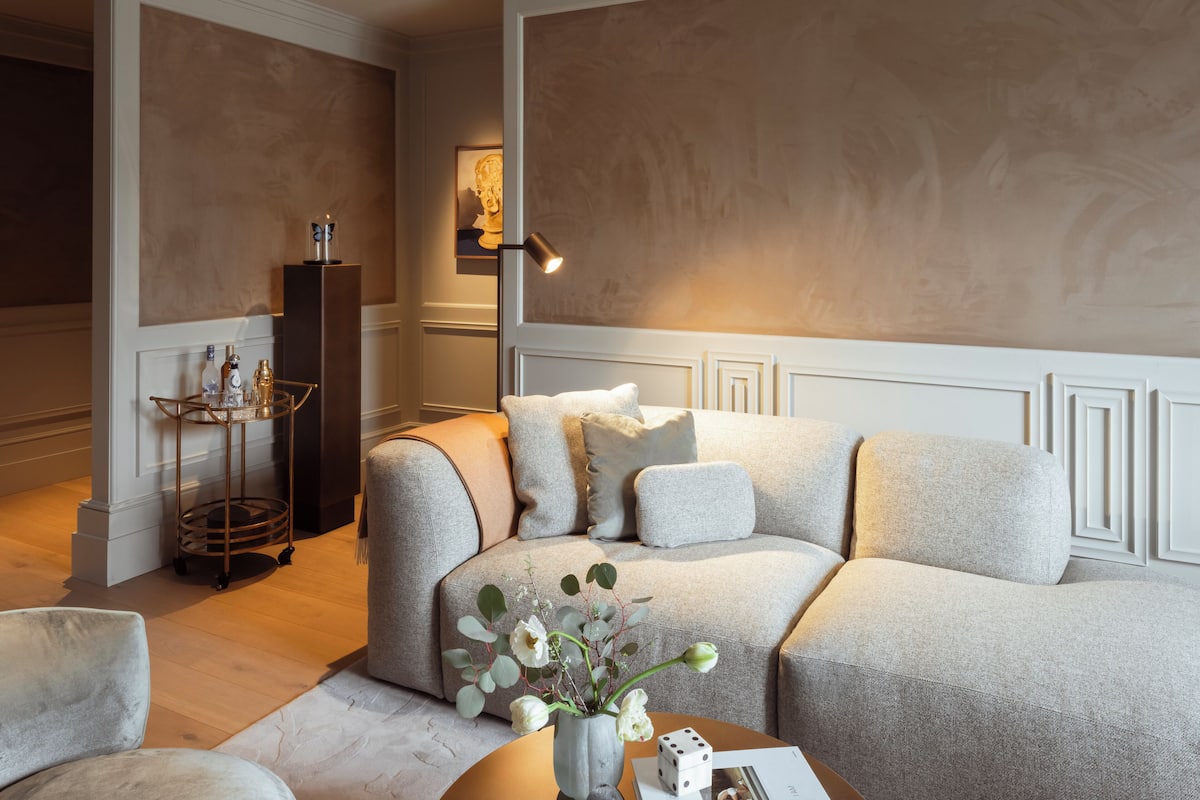 A modern living room with a sectional sofa, a small round table with a vase of flowers, and a decorative lamp.