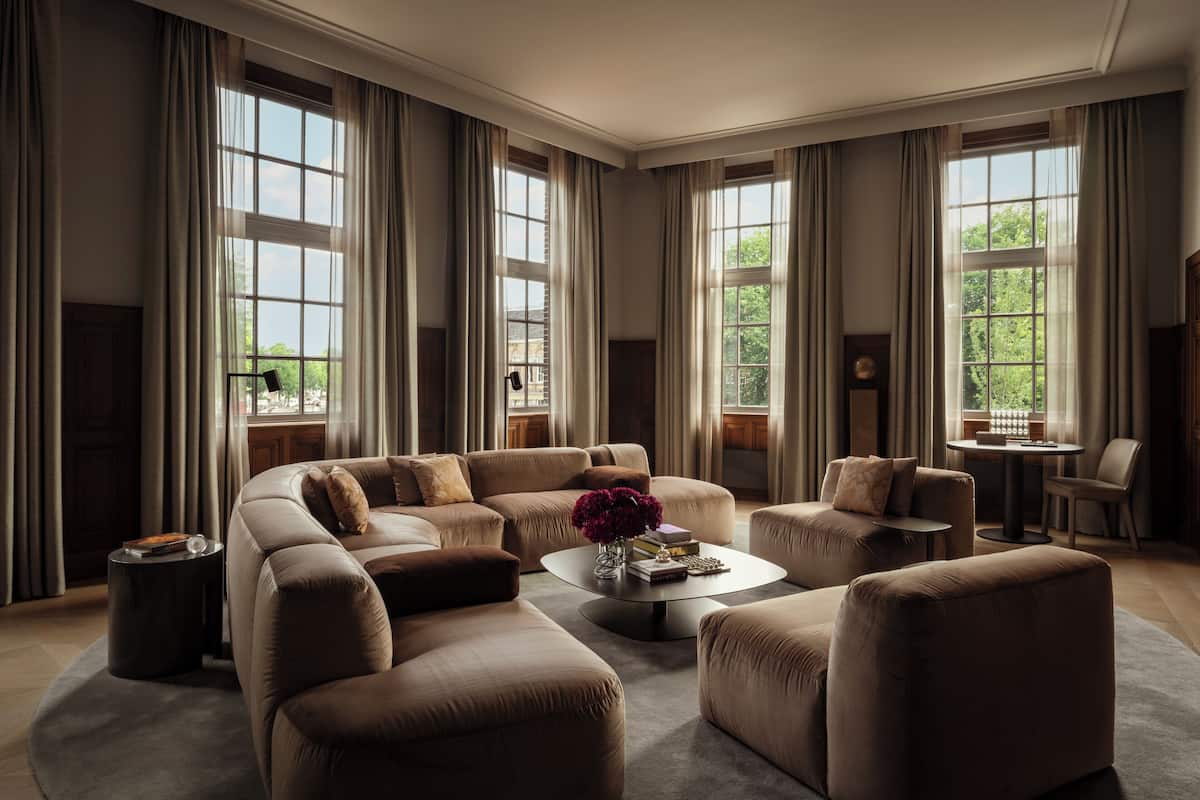 A spacious living room with a large sectional sofa, a coffee table, and a small side table. There are large windows with curtains, a fireplace, and a view of greenery outside.