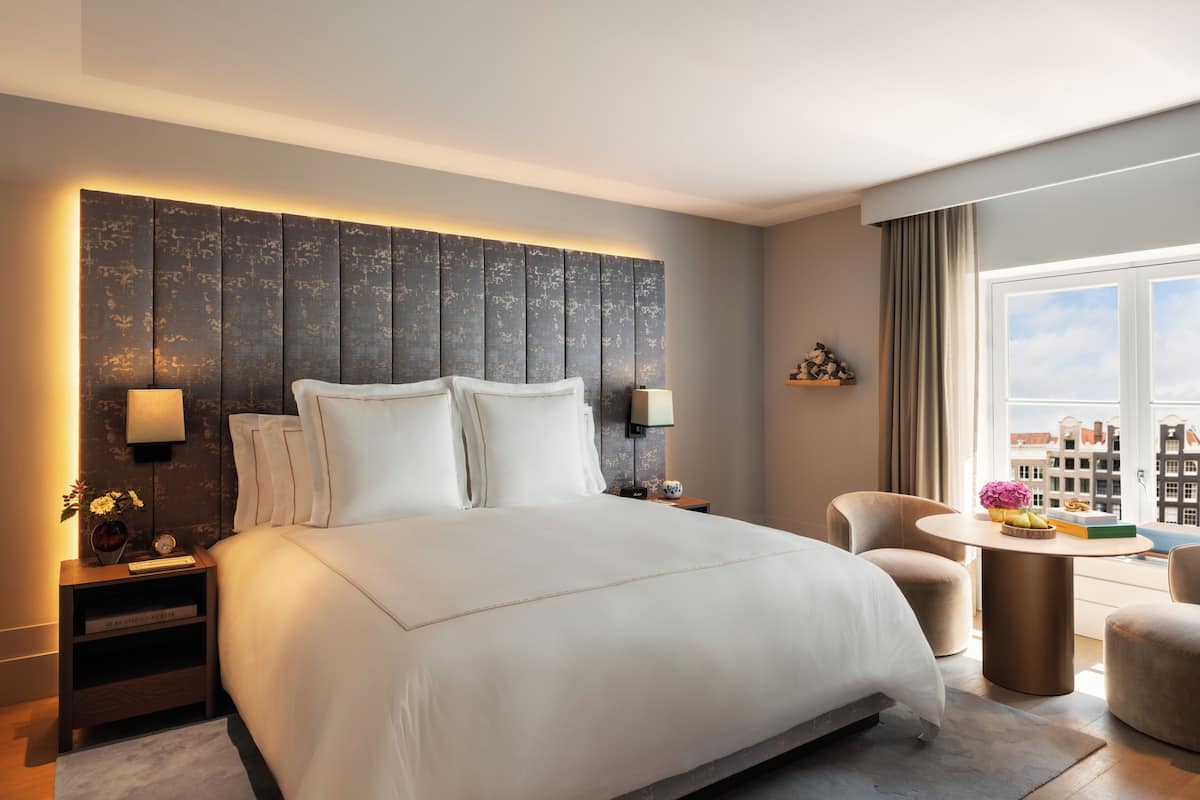 A modern hotel room with a large bed, bedside lamps, a small table with flowers, and a view of buildings through the window.