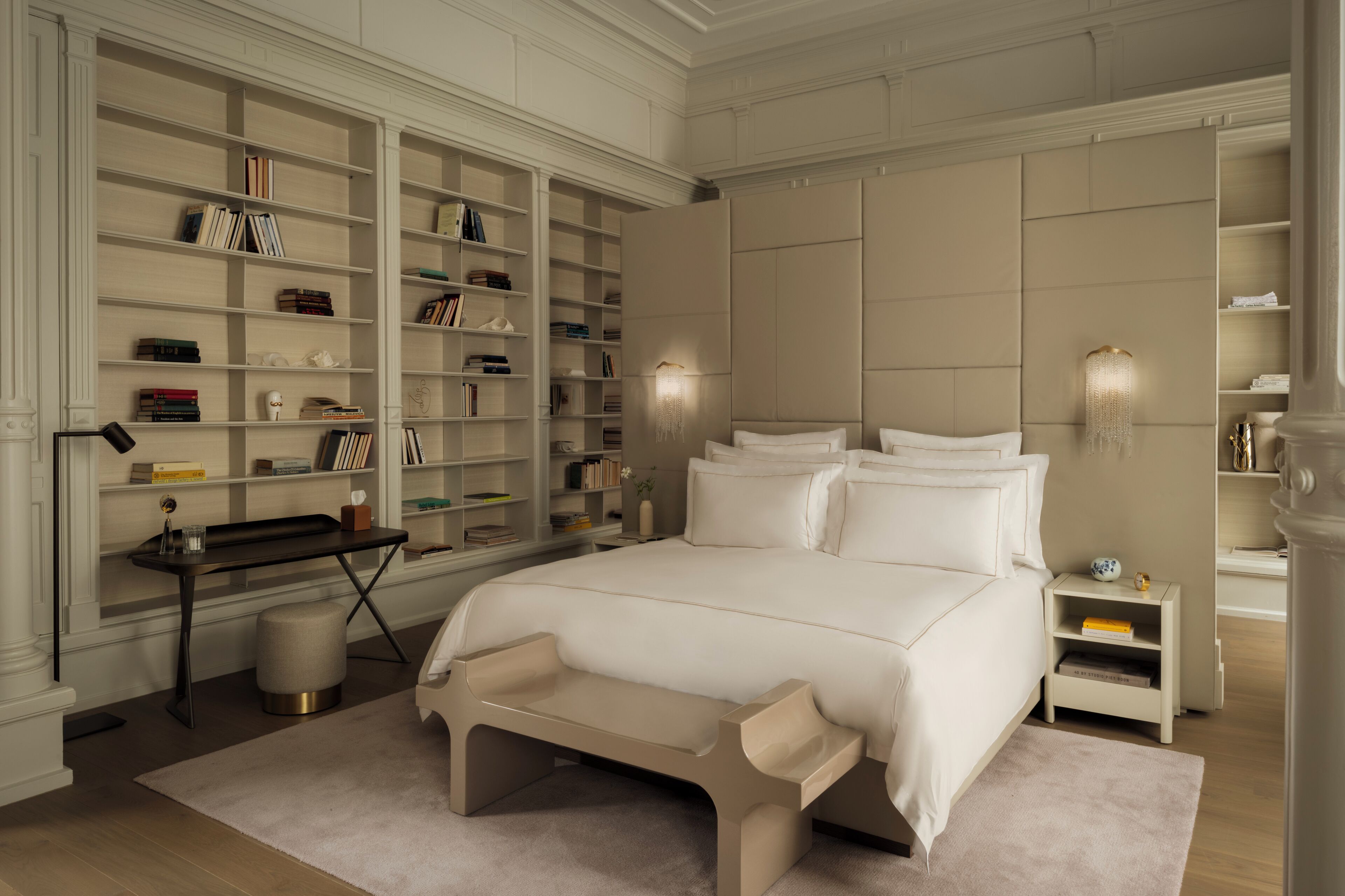 A bedroom with a large bed, a bedside table, a nightstand, a bookshelf, and a desk with a lamp.