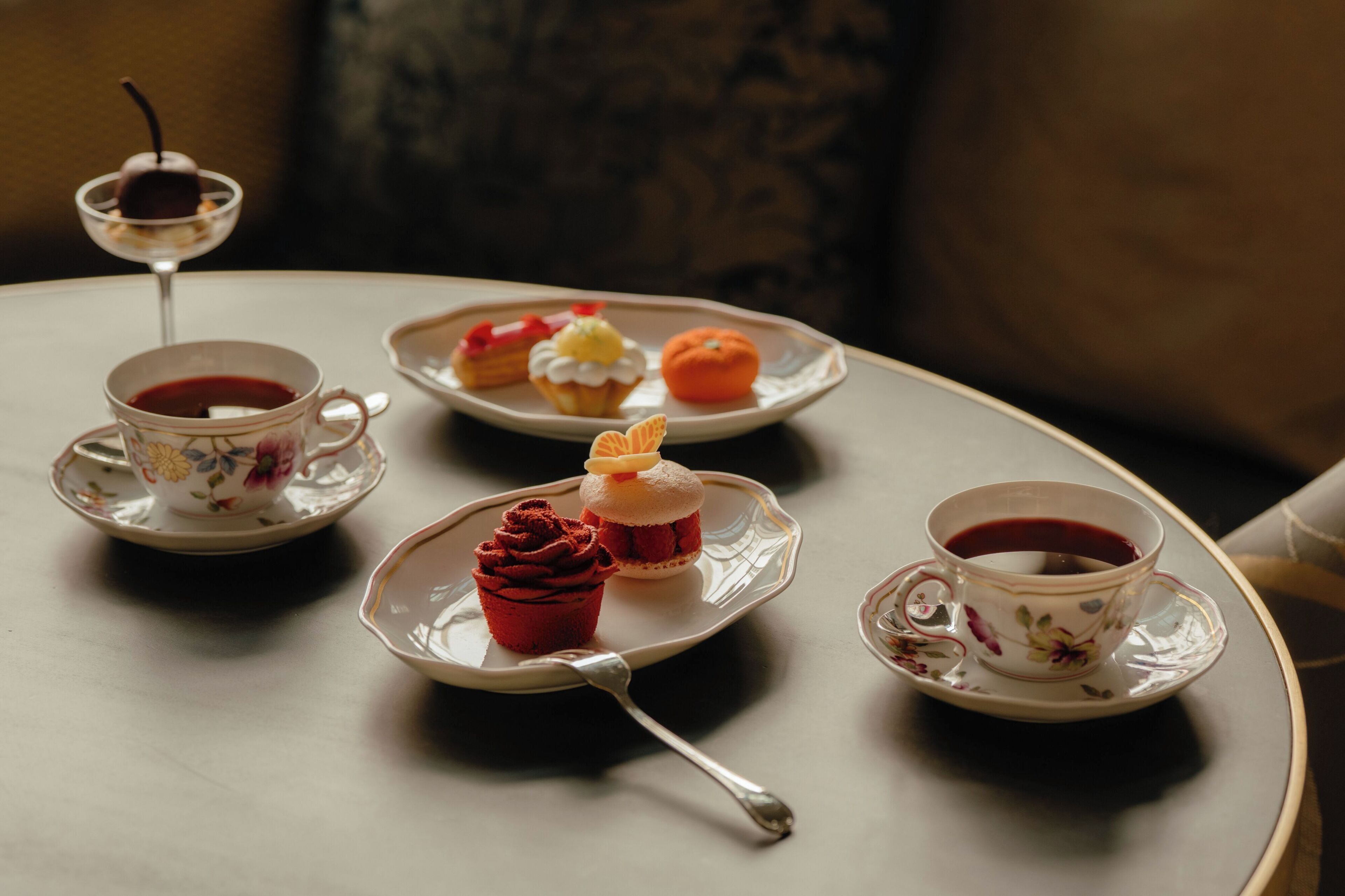 A table with a floral-patterned cup and saucer, a plate with a cupcake and a small dessert, and a glass of red beverage.