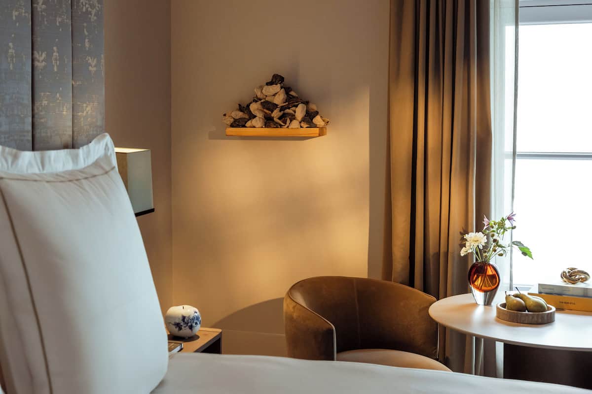 A hotel room with a bed, a chair, a small table with a vase and pears, and a shelf with dried flowers.