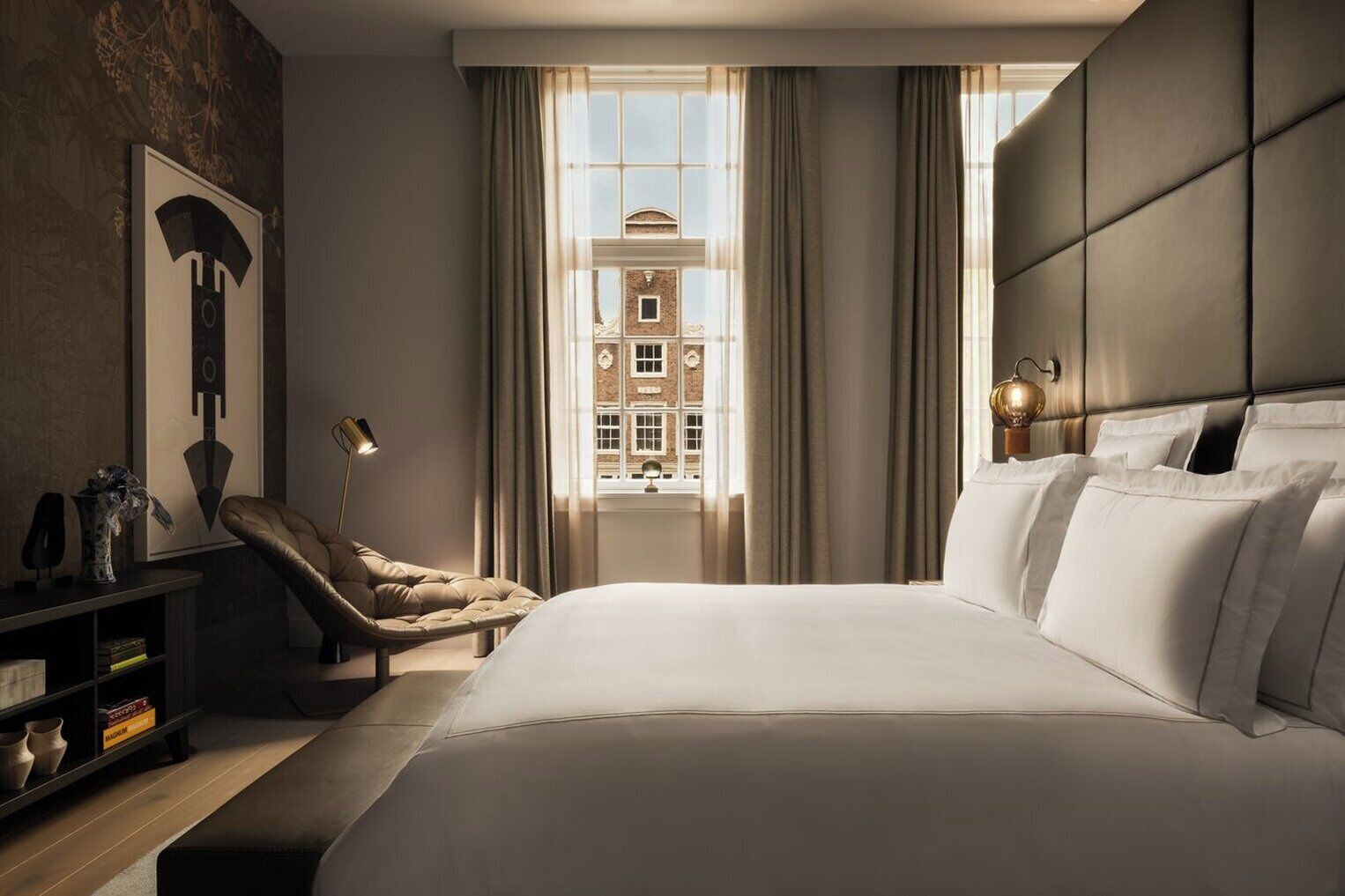 A hotel room with a large bed, a chair, a desk, and a view of a building through the window.