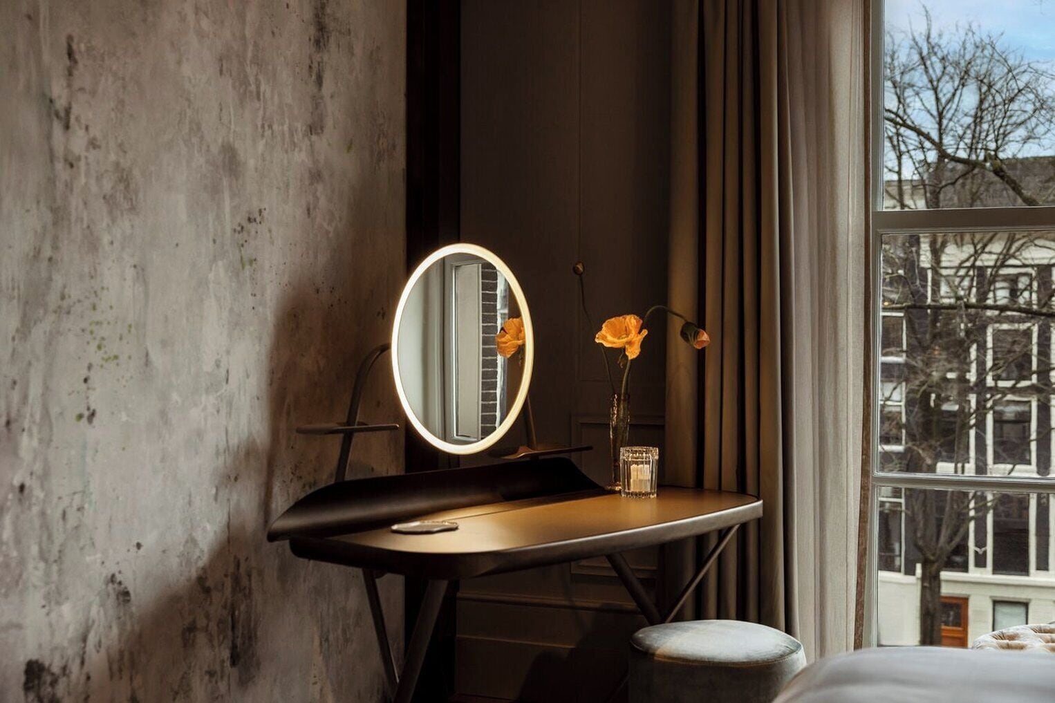 Una habitación con cama, tocador con espejo, un florero con flores y ventana con vista a edificios.