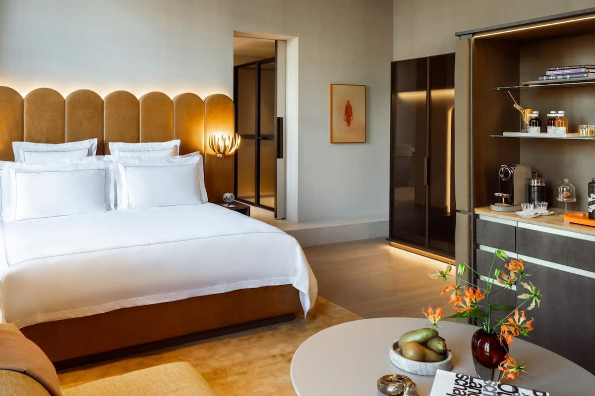 A modern hotel room with a large bed, bedside lamps, a minibar, and a view of the hallway.