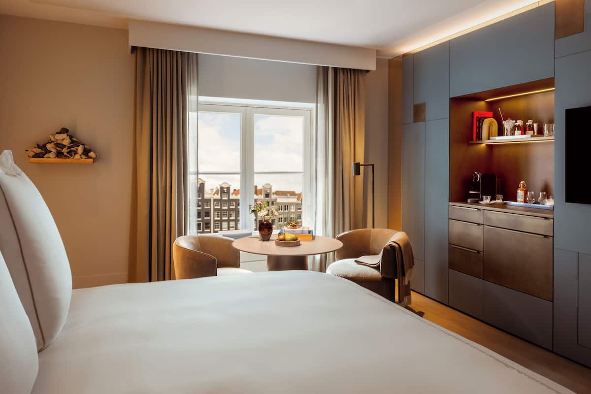 A hotel room with a bed, a round table with two chairs, a small table with a vase, a minibar, and a large window with curtains.