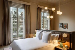 Hotellihuone, jossa on suuri sänky, kaksi yöpöytää, pieni sivupöytä, peili ja ikkunoista avautuva näkymä rakennuksiin.