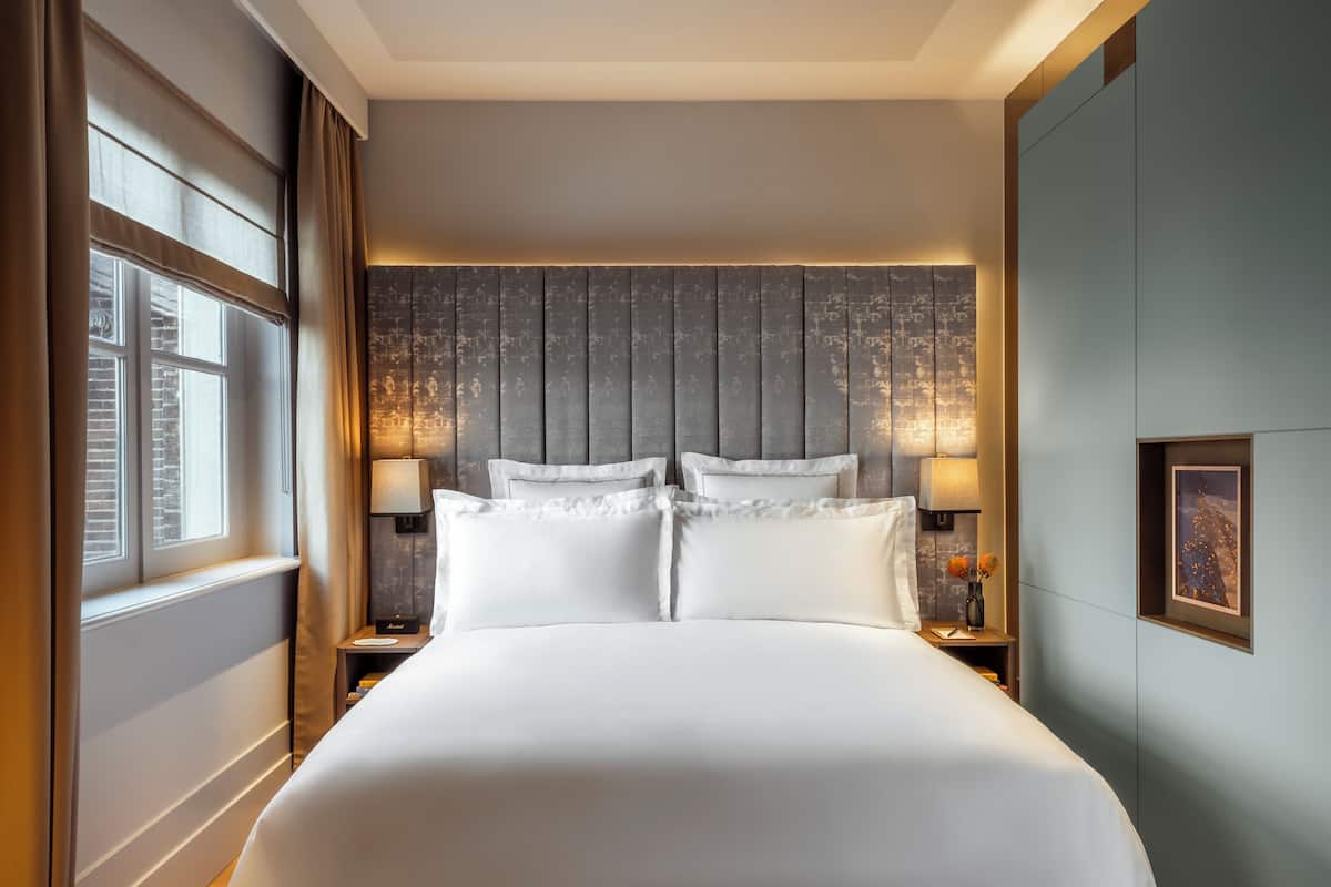 A neatly made bed with white linens in a modern room with a wooden headboard, two bedside lamps, and a framed picture on the wall.