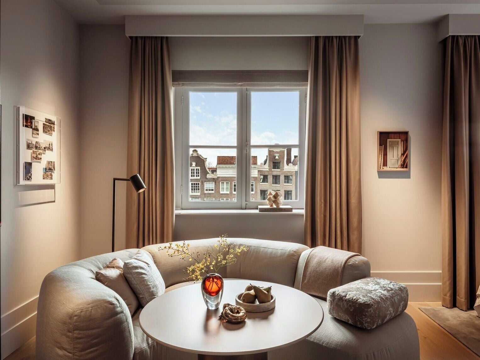 Een moderne woonkamer met een ronde tafel, een beige bank en uitzicht op gebouwen door een raam.
