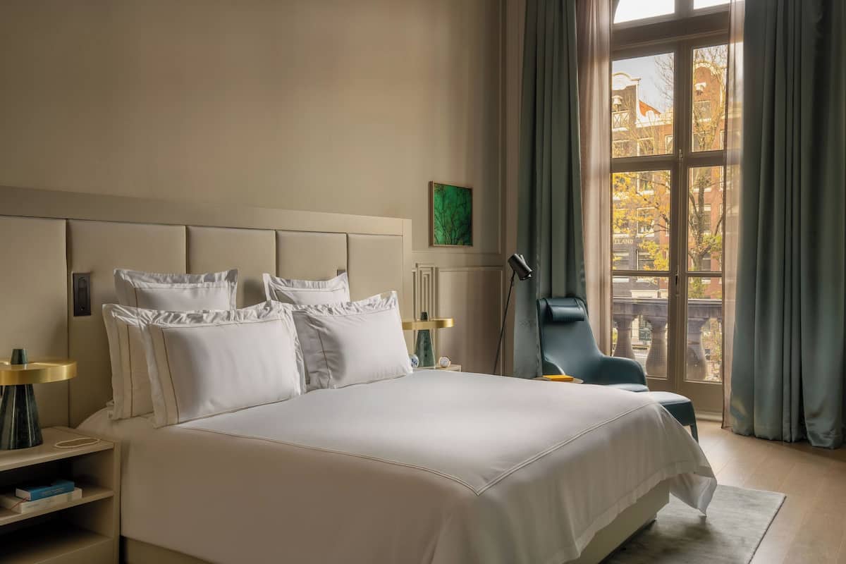 A hotel room with a large bed, a chair, a nightstand, a lamp, and a window with curtains.