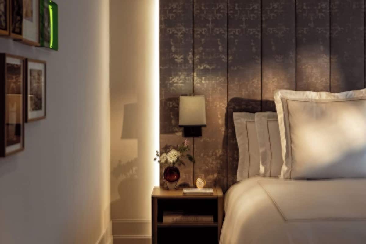 A bedroom with a wooden headboard, a bedside table with a lamp, a vase with flowers, and a clock.