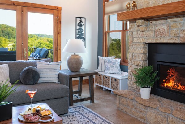 A cozy living room with a fireplace, a sofa, a coffee table with snacks, and a view of the outdoors.