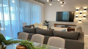 Una sala de estar moderna con una mesa de comedor de vidrio, sofás grises, un televisor de pantalla plana y un aplique de pared con diseño geométrico.