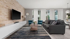 A modern living room with a flat-screen TV mounted on a brick wall, a grey sofa, two blue armchairs, a wooden coffee table, and a striped rug. - Incredible 4BD home with Pool and BBQ near Disney (Kissimmee)